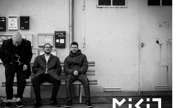 Drei M&auml;nner sitzen auf einer Bank vor einer Wand mit Briefk&auml;sten. Einer steht auf, w&auml;hrend die anderen sitzen. Der Schriftzug 'MIKIO' ist sichtbar., &copy; Foto MIKIO