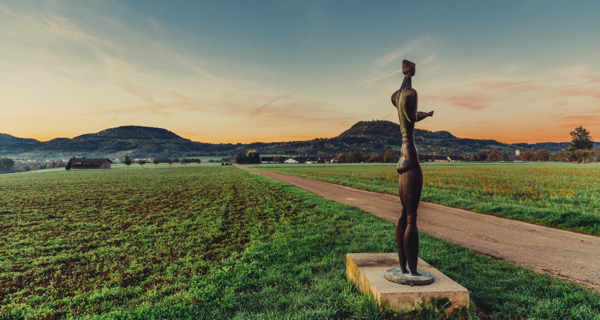 Eine Skulptur steht auf einem Feldweg, umgeben von grünen Feldern und Hügeln im Hintergrund, während die Sonne untergeht., © Landratsamt Ostalbkreis