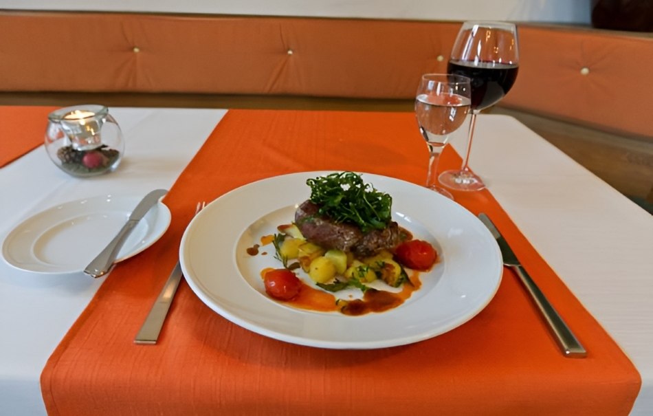 Ein elegant gedeckter Tisch mit einem Teller Fleischgericht, Gemüse und Soße. Daneben stehen ein Glas Rotwein und Wasser. Die Tischdecke ist orange-weiß., © Hotel Neuwirtshaus