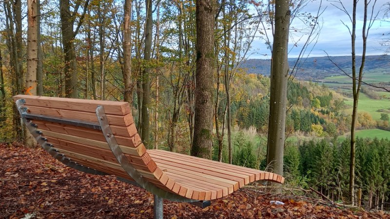 Holzliege im Wald am Lehberg, mit Blick auf eine herbstliche, hügelige Landschaft und grüne Felder im Hintergrund., © Stadt Gaildorf Holzliege im Wald am Lehberg, mit Blick auf eine herbstliche, hügelige Landschaft und grüne Felder im Hintergrund., © Stadt Gaildorf