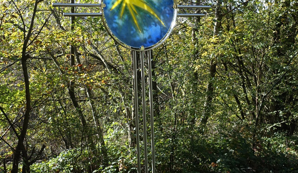 Ein Kreuz mit einem blauen und gelben Glasornament steht in einem Wald, umgeben von Bäumen und Pflanzen., © Foto: Cornelia Steinbach