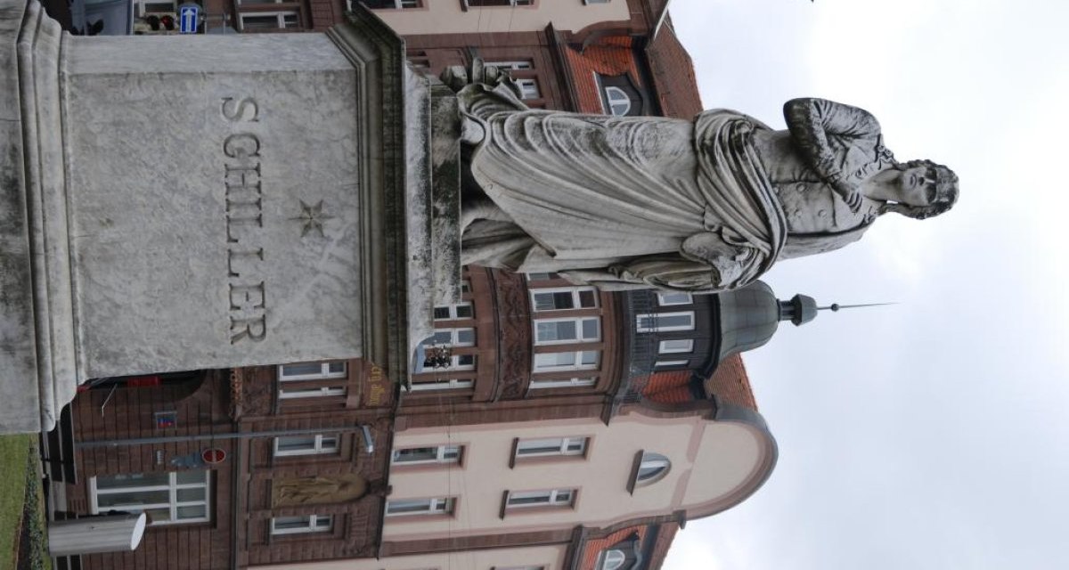 Das Schillerdenkmal steht vor einem historischen Gebäude. Die Statue zeigt eine Person in klassischer Pose auf einem Sockel mit der Aufschrift 'Schiller'., © Ludwigsburg - Stuttgart-Marketing GmbH Das Schillerdenkmal steht vor einem historischen Gebäude. Die Statue zeigt eine Person in klassischer Pose auf einem Sockel mit der Aufschrift 'Schiller'., © Ludwigsburg - Stuttgart-Marketing GmbH