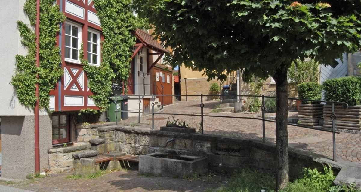 Fachwerkhaus mit Efeu bewachsen, daneben ein Baum und ein kleiner Brunnen in einer gepflasterten Gasse., &copy; Natur.Nah. Sch&ouml;nbuch & Heckeng&auml;u