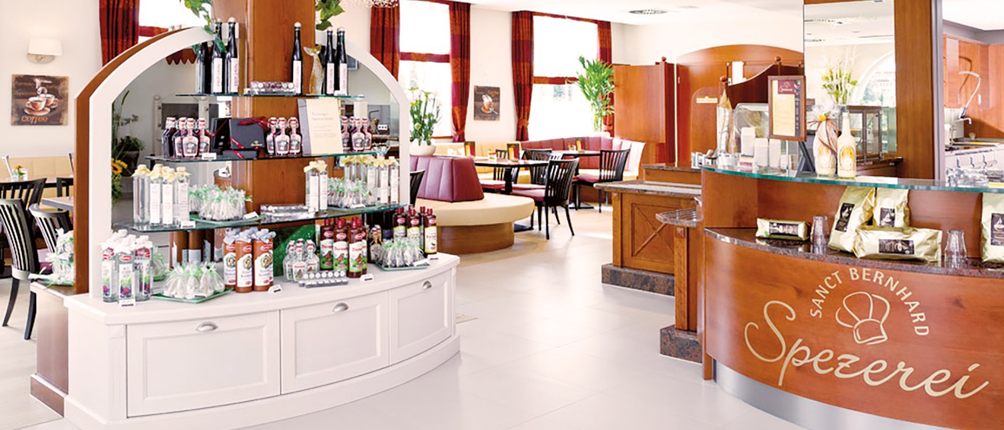Ein gemütliches Kaffeehaus mit Regalen voller Produkte und einem einladenden Sitzbereich. Holzmöbel und helle Dekoration dominieren den Raum., © Kräuterhaus Sanct Bernhard KG