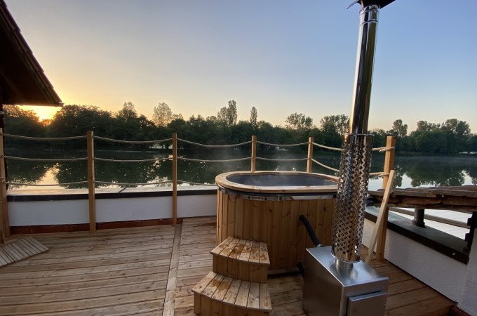 Holzterrasse mit Hot Tub, daneben ein Ofen. Im Hintergrund ein Fluss und Bäume bei Sonnenuntergang., © Cool-Tours StattReisen Holzterrasse mit Hot Tub, daneben ein Ofen. Im Hintergrund ein Fluss und Bäume bei Sonnenuntergang., © Cool-Tours StattReisen