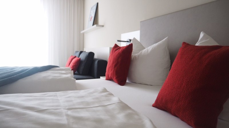 Modernes Hotelzimmer mit weißen Bettlaken, roten Kissen und einem Sofa. Ein Bild hängt an der Wand, helle Vorhänge lassen Licht herein., © TOMAS