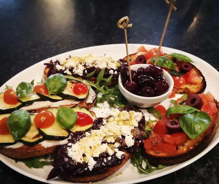 Ein Teller mit Antipasti: Bruschetta mit Tomaten, Zucchini, Oliven und Basilikum, dazu eine Schale mit Oliven in der Mitte., © La buona Forketta Ein Teller mit Antipasti: Bruschetta mit Tomaten, Zucchini, Oliven und Basilikum, dazu eine Schale mit Oliven in der Mitte., © La buona Forketta