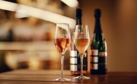 Zwei Weingl&auml;ser mit ros&eacute;farbenem Getr&auml;nk stehen auf einem Holztisch, im Hintergrund sind unscharf Flaschen zu sehen., &copy; Sommer.Auszeit. Foto: Teamwerk