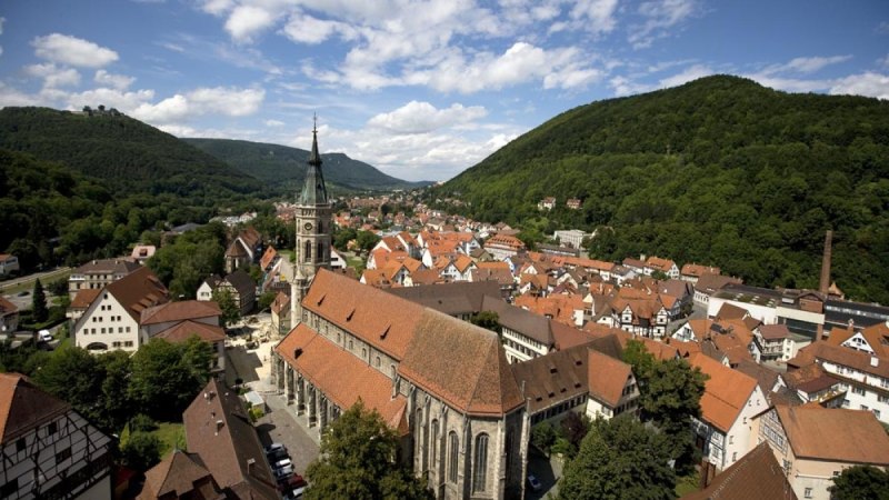 Luftaufnahme der Kirche St. Amandus in Bad Urach, umgeben von roten D&auml;chern und gr&uuml;nen H&uuml;geln unter blauem Himmel mit Wolken., &copy; Bad Urach Tourismus