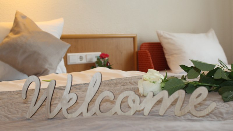 Ein Bett mit Kissen, einer wei&szlig;en Rose und einem 'Welcome'-Schild. Im Hintergrund eine Steckdose und ein roter Sessel., &copy; TOMAS