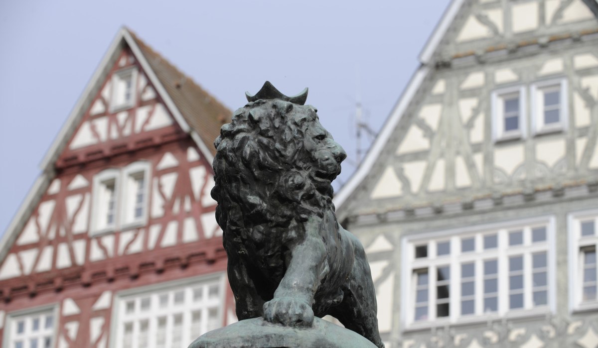 Eine Bronzestatue eines Löwen mit Krone steht vor zwei Fachwerkhäusern. Die Häuser haben weiße und rote sowie weiße und graue Fassaden., © Land der 1000 Hügel - Kraichgau-Stromberg