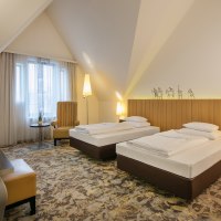 Ein modernes Hotelzimmer mit zwei Einzelbetten, einem Schreibtisch, einem Sessel und einem Flachbildfernseher. Die Einrichtung ist in warmen Tönen gehalten., © ARCOTEL Hotels
