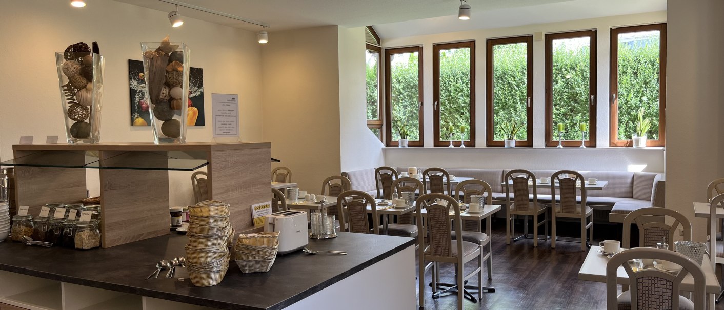 Heller Frühstücksraum mit gedeckten Tischen, großen Fenstern und einem Buffetbereich. Dekorative Vasen und Pflanzen schmücken den Raum., © TOMAS