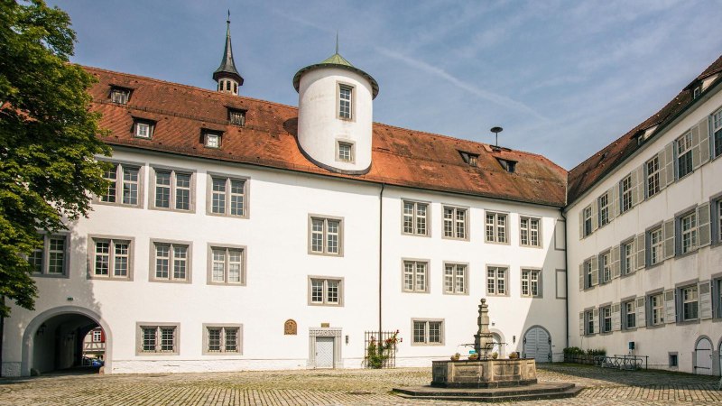 Der Innenhof von Schloss Waldenbuch zeigt weiße Gebäude mit roten Dächern und einen zentralen Brunnen. Ein blauer Himmel rundet die Szene ab., © SMG, Sarah Schmid Der Innenhof von Schloss Waldenbuch zeigt weiße Gebäude mit roten Dächern und einen zentralen Brunnen. Ein blauer Himmel rundet die Szene ab., © SMG, Sarah Schmid
