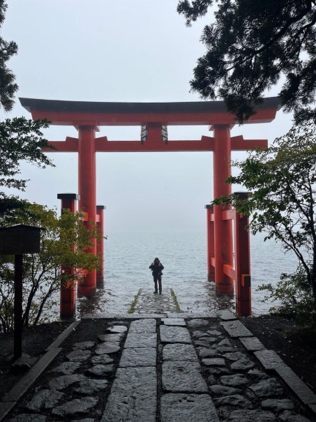 Ein rotes Torii-Tor ragt aus dem Wasser, w&auml;hrend eine Person davor steht. Der neblige Himmel und der steinige Weg schaffen eine mystische Atmosph&auml;re., &copy; Daniel Hikel
