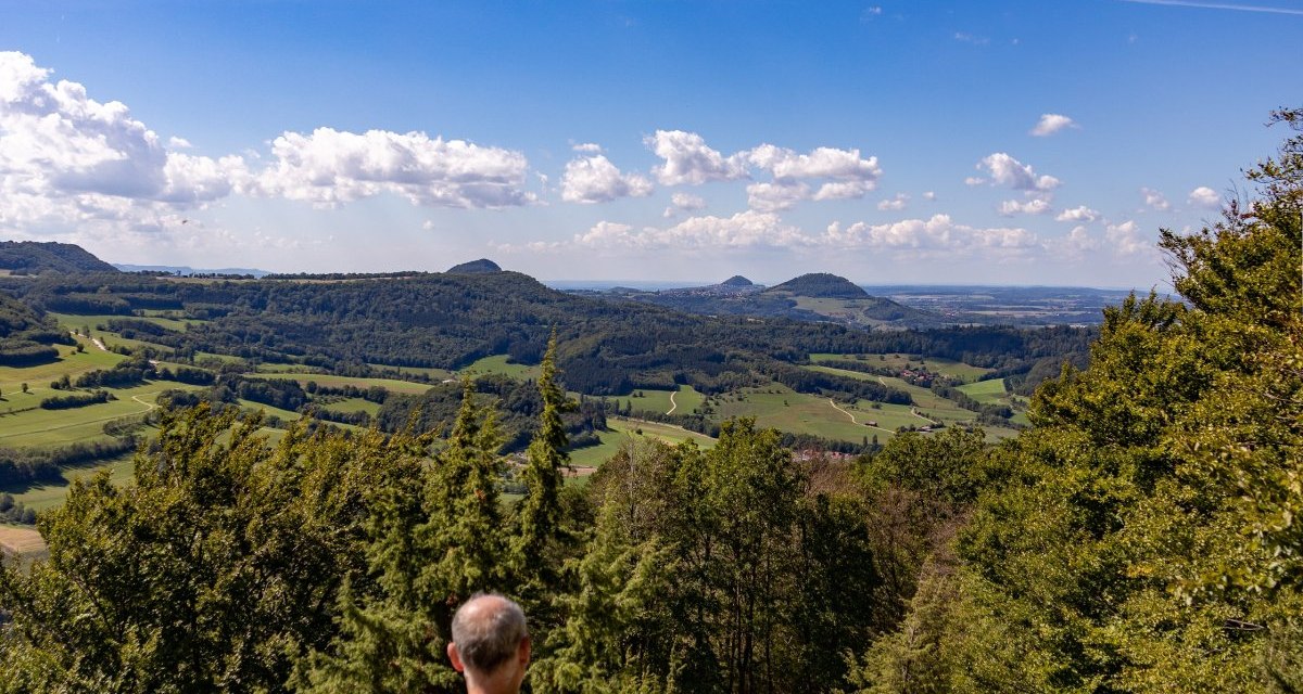 Eine Person sitzt auf einer Bank und blickt auf eine weite, grüne Landschaft unter einem klaren, blauen Himmel mit wenigen Wolken., © Foto Thomas Zehnder Eine Person sitzt auf einer Bank und blickt auf eine weite, grüne Landschaft unter einem klaren, blauen Himmel mit wenigen Wolken., © Foto Thomas Zehnder