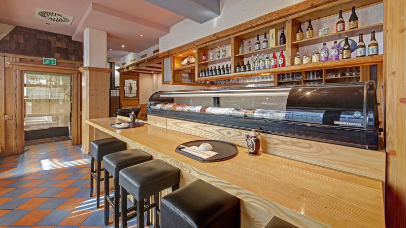 Ein japanisches Restaurant mit Sushi-Theke, Barhockern und Regalen voller Sake-Flaschen. Holzelemente dominieren das Interieur., &copy; Restaurant Kurose