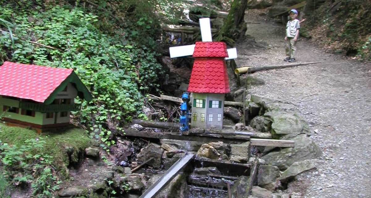 Eine Miniatur-Wassermühle mit rotem Dach im Wald. Ein Kind steht auf einem Pfad daneben. Umgeben von Bäumen und grünem Laub., © Remstal Tourismus e.V. Eine Miniatur-Wassermühle mit rotem Dach im Wald. Ein Kind steht auf einem Pfad daneben. Umgeben von Bäumen und grünem Laub., © Remstal Tourismus e.V.