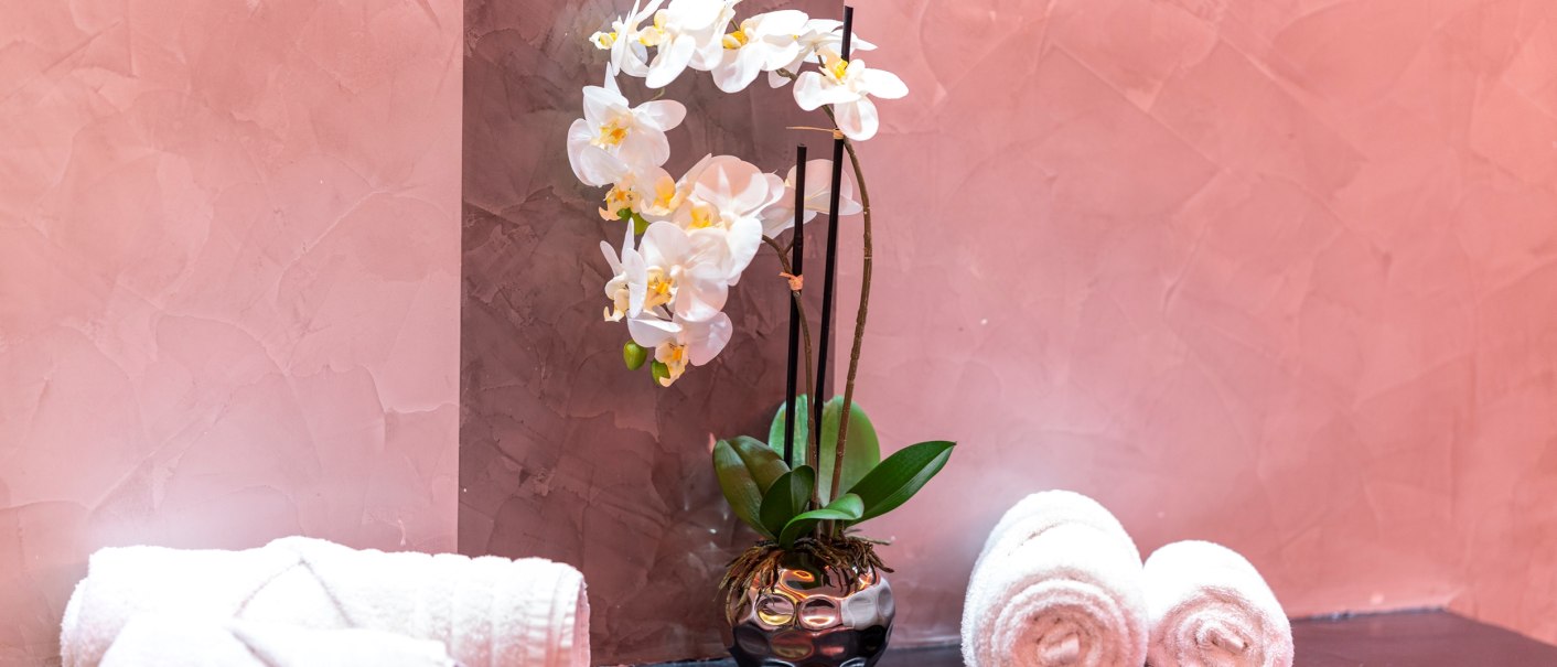 Weiße Orchidee in einer glänzenden Vase, umgeben von gerollten weißen Handtüchern auf einem dunklen Tisch vor einer rosa Wand., © Plaza Residence Stuttgart-Ditzingen Weiße Orchidee in einer glänzenden Vase, umgeben von gerollten weißen Handtüchern auf einem dunklen Tisch vor einer rosa Wand., © Plaza Residence Stuttgart-Ditzingen