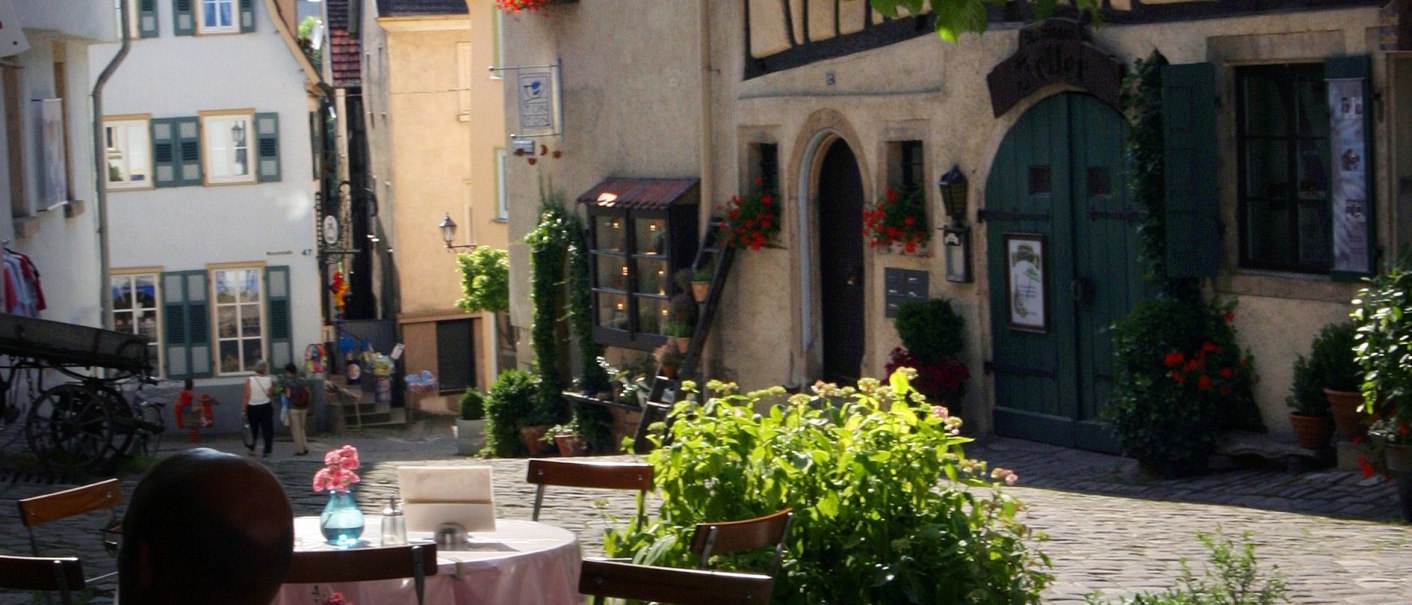 Gemütliches Café in einer Altstadt mit Fachwerkhäusern, blühenden Blumen und einem Gast im Vordergrund. Die Szene wirkt idyllisch und einladend., © Nürtingen Gemütliches Café in einer Altstadt mit Fachwerkhäusern, blühenden Blumen und einem Gast im Vordergrund. Die Szene wirkt idyllisch und einladend., © Nürtingen