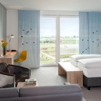 Modernes Hotelzimmer mit großem Bett, Schreibtisch, Sessel und Balkon mit Aussicht auf Landschaft. Dekor mit blauen Vorhängen und Vogelmotiven., © Essential by Dorint