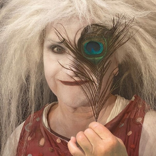 Eine Person mit wildem, weißem Haar und weiß geschminktem Gesicht hält eine Pfauenfeder vor einem Auge. Sie trägt ein rotes, gepunktetes Oberteil., © Mauerwerk GmbH Eine Person mit wildem, weißem Haar und weiß geschminktem Gesicht hält eine Pfauenfeder vor einem Auge. Sie trägt ein rotes, gepunktetes Oberteil., © Mauerwerk GmbH