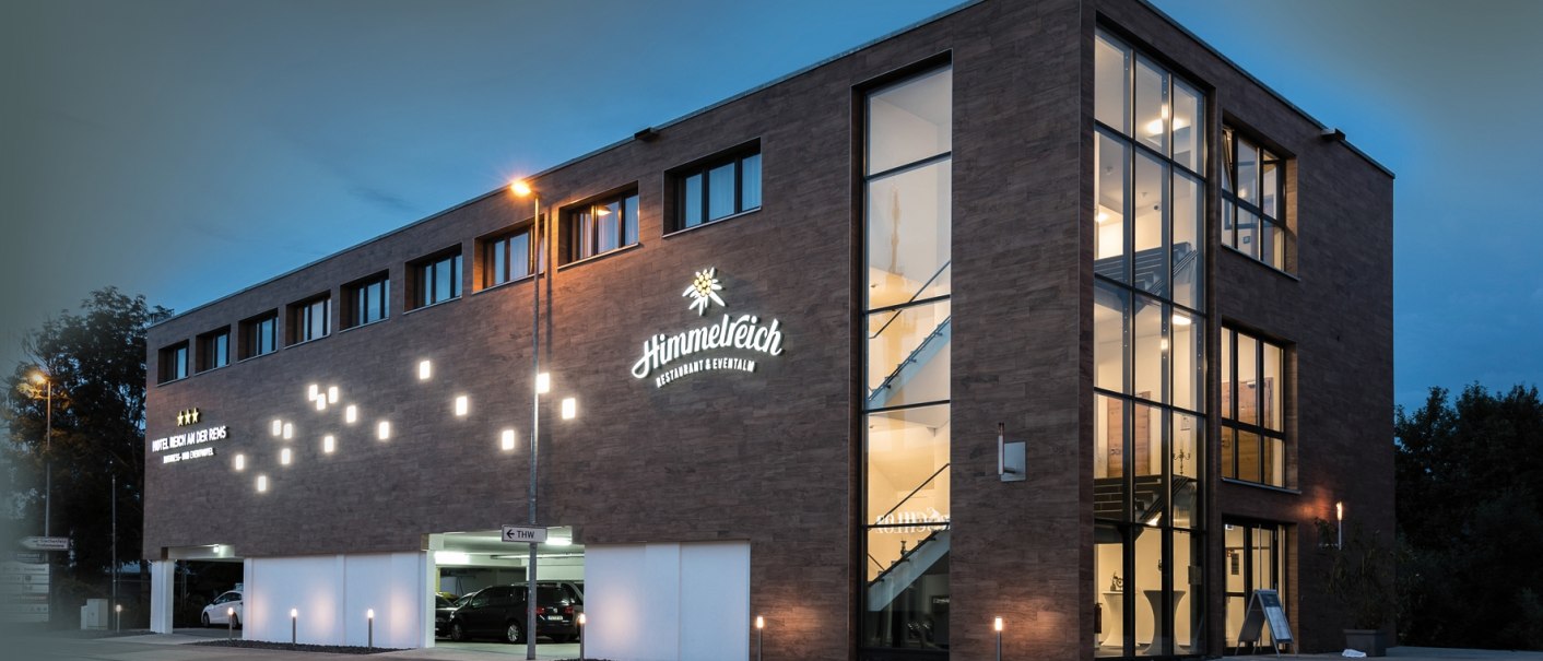 Modernes Hotelgebäude bei Nacht, beleuchtet mit großen Fenstern und Schildern. "Himmelreich Restaurant & Eventlocation" ist sichtbar., © Hotel Reich an der Rems