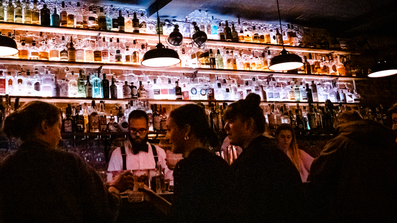Eine stimmungsvolle Bar mit beleuchteten Flaschenregalen. Menschen unterhalten sich, während ein Barkeeper Getränke zubereitet., © Stuttgart-Marketing GmbH, Sarah Schmid