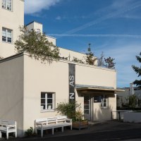 Beige Architekturgalerie mit Flachdach, Bänken und großem Nadelbaum vor blauem Himmel., © Thomas Fütterer Beige Architekturgalerie mit Flachdach, Bänken und großem Nadelbaum vor blauem Himmel., © Thomas Fütterer
