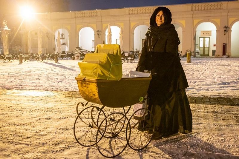 Eine Person in schwarzem Mantel steht mit einem antiken Kinderwagen im Schnee vor einem beleuchteten Gebäude mit Bögen., © Tourist Information und Ticket Service Ludwigsburg