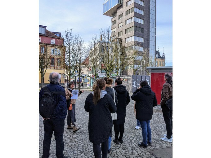 Eine Gruppe von Menschen betrachtet ein modernes Hochhaus auf einem gepflasterten Platz. Im Hintergrund sind weitere Gebäude und Bäume zu sehen., © Eigenbetrieb Tourismus und Citymanagement Schorndorf