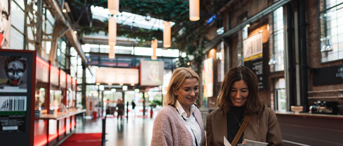 Zwei Frauen stehen im Foyer eines Theaterhauses und schauen auf Tickets. Im Hintergrund sind Poster und eine moderne Architektur zu sehen., © Stuttgart-Marketing GmbH, wpsteinheisser