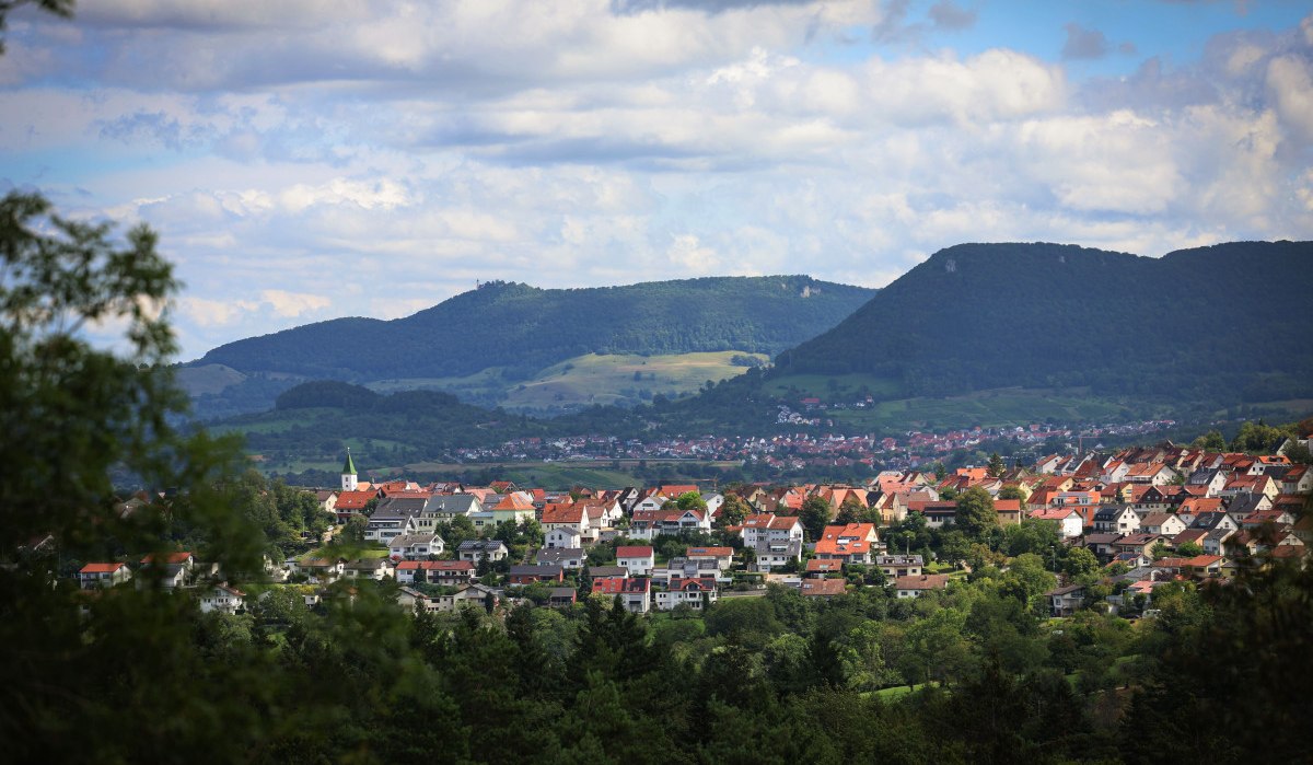 Kleine Stadt mit roten Dächern, umgeben von grünen Hügeln und Wäldern. Bewölkter Himmel im Hintergrund.