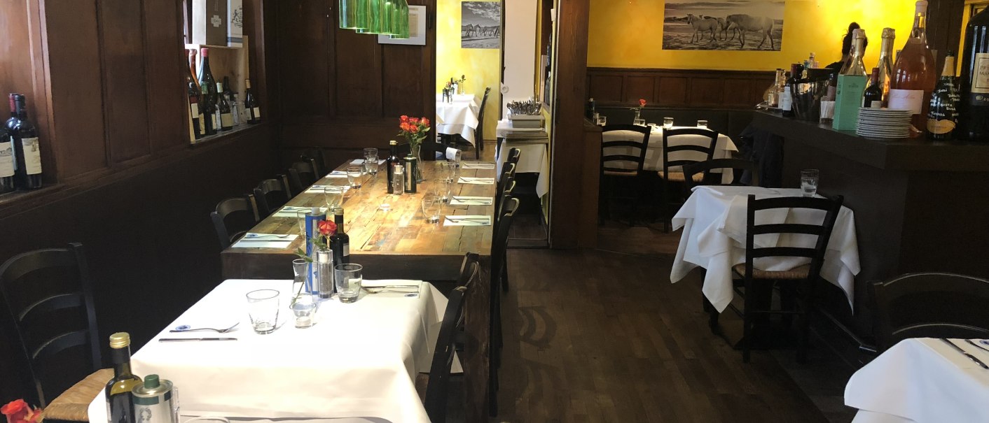 Gemütliches Restaurant mit Holztischen, Weinflaschen an den Wänden und gelben Wänden. Tische sind mit weißen Tischdecken gedeckt., © Antipasto Wein und mehr
