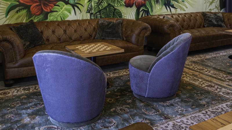 Gemütliche Sitzecke mit braunen Ledersofas, blauen Sesseln und einem Schachbrett-Tisch vor einer Wand mit tropischem Pflanzenmuster., © 13 6 Design GmbH (Judenhof 10, 89073 Ulm) Gemütliche Sitzecke mit braunen Ledersofas, blauen Sesseln und einem Schachbrett-Tisch vor einer Wand mit tropischem Pflanzenmuster., © 13 6 Design GmbH (Judenhof 10, 89073 Ulm)