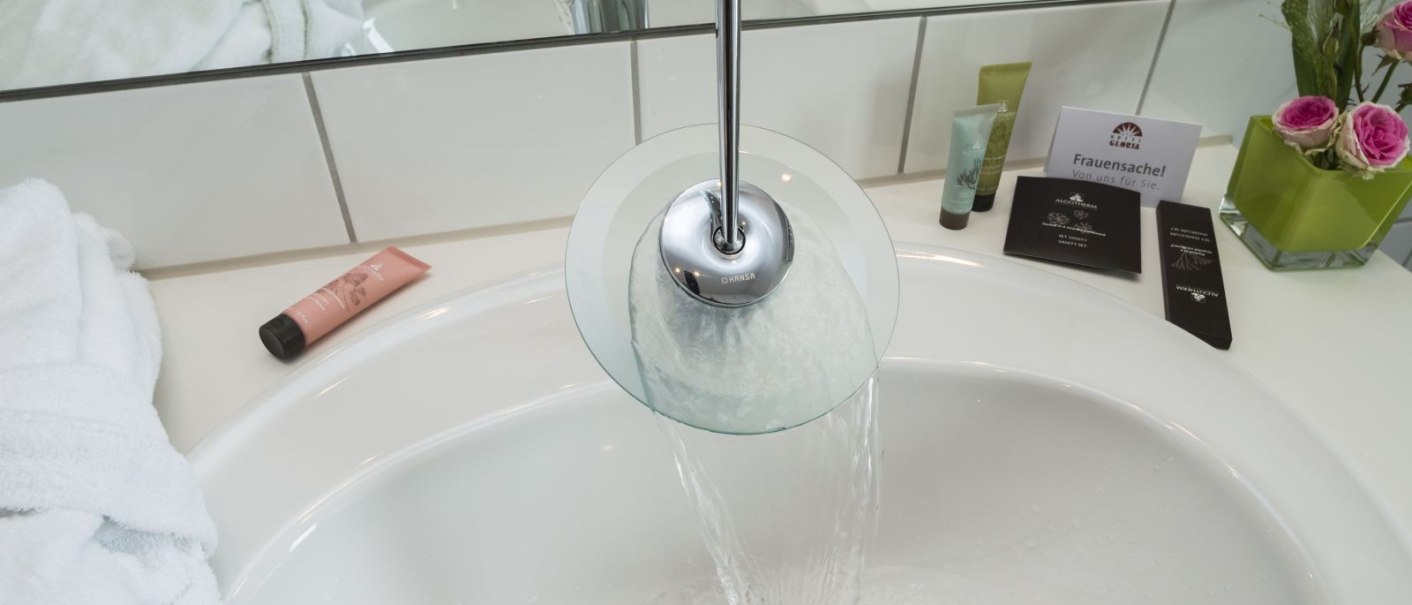 Modernes Badezimmer mit fließendem Wasser im Waschbecken, Handtuch, Pflegeprodukten und Blumen., © Hotel Gloria GmbH Restaurant Möhringer Hexle