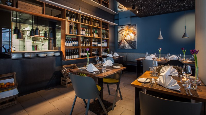Stilvolles Restaurant mit gedeckten Tischen, Weinregalen und moderner Beleuchtung. Eine offene Küche und ein Kunstwerk an der Wand sind sichtbar., © Belser Stilvolles Restaurant mit gedeckten Tischen, Weinregalen und moderner Beleuchtung. Eine offene Küche und ein Kunstwerk an der Wand sind sichtbar., © Belser
