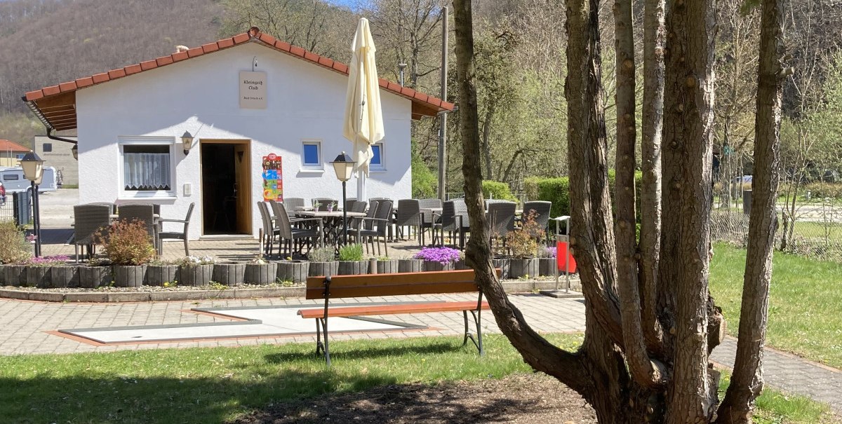 Ein Minigolfplatz mit einem kleinen weißen Gebäude und Sitzbereich im Freien, umgeben von Bäumen und Natur.