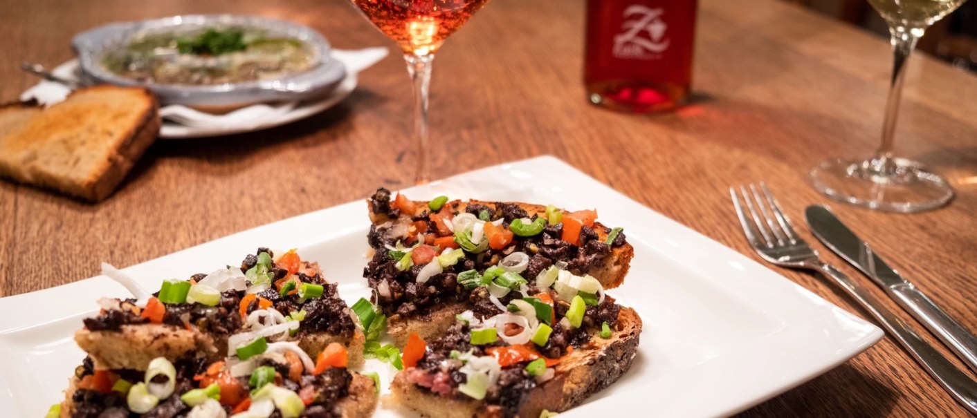 Bruschetta mit Olivenpaste und Gemüse auf einem weißen Teller, daneben ein Glas Roséwein auf einem Holztisch., © Weingut Zaiß
