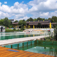 Das Naturfreibad Herrenberg zeigt einen Pool mit Holzdeck, Felsenwand und umgebender grüner Landschaft unter blauem Himmel mit Wolken., © Stadtwerke Herrenberg Das Naturfreibad Herrenberg zeigt einen Pool mit Holzdeck, Felsenwand und umgebender grüner Landschaft unter blauem Himmel mit Wolken., © Stadtwerke Herrenberg