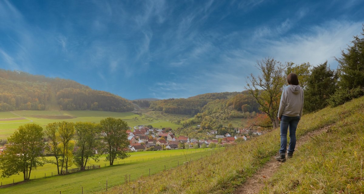 Eine Person steht auf einem Hügel und blickt auf ein Dorf in einem Tal, umgeben von grünen Wiesen und bewaldeten Hügeln unter einem blauen Himmel., © Foto: Mario Klaiber Eine Person steht auf einem Hügel und blickt auf ein Dorf in einem Tal, umgeben von grünen Wiesen und bewaldeten Hügeln unter einem blauen Himmel., © Foto: Mario Klaiber