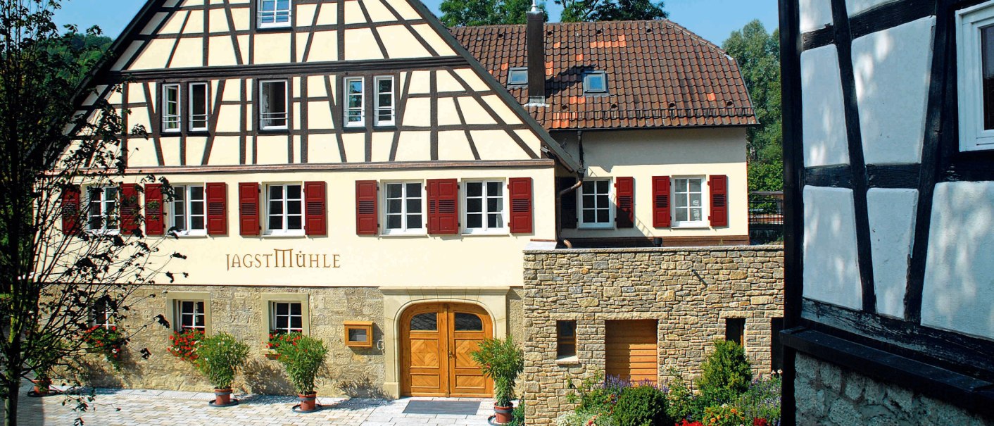 Fachwerkhaus mit rotem Dach und Fensterläden, beschriftet mit 'Jagstmühle'. Vor dem Haus stehen Pflanzen in Töpfen., © Landgasthof + Hotel Jagstmühle Fachwerkhaus mit rotem Dach und Fensterläden, beschriftet mit 'Jagstmühle'. Vor dem Haus stehen Pflanzen in Töpfen., © Landgasthof + Hotel Jagstmühle