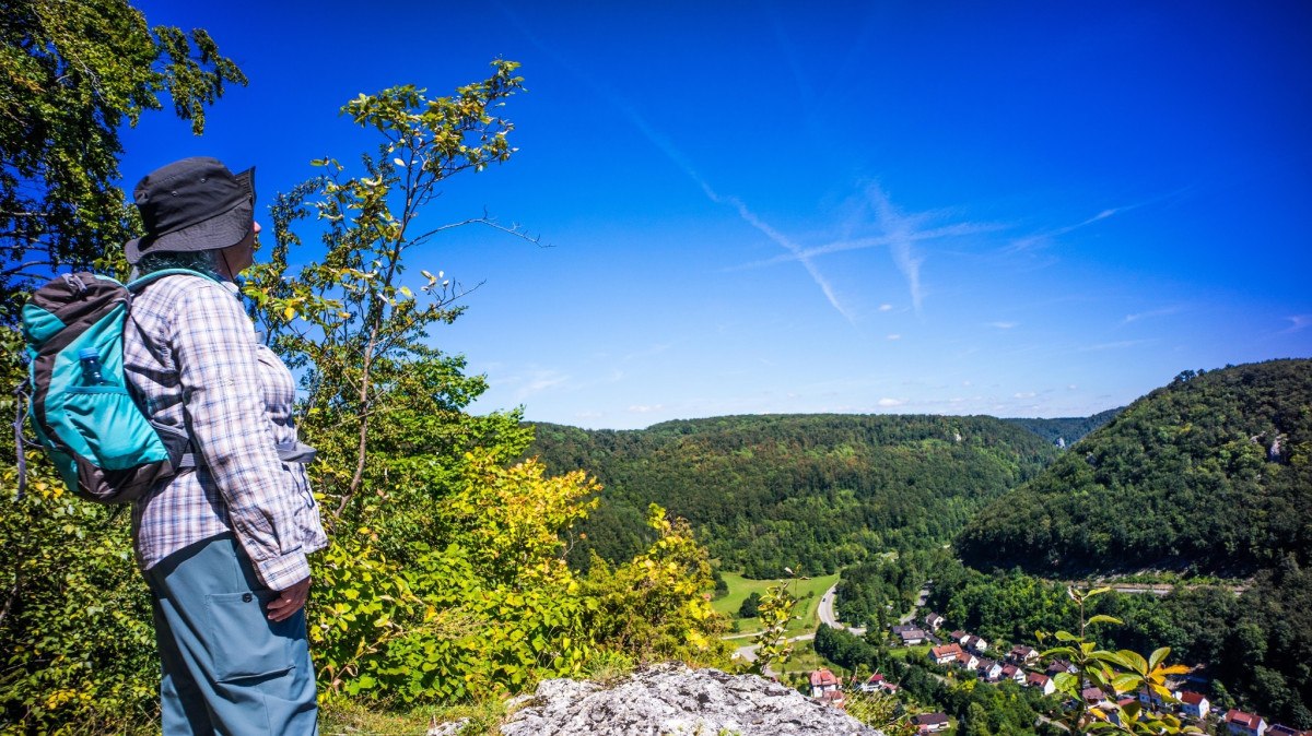 Eine Person mit Rucksack steht auf einem Hügel und blickt auf ein bewaldetes Tal mit einem Dorf. Der Himmel ist klar und blau., © Stadt Geislingen an der Steige Eine Person mit Rucksack steht auf einem Hügel und blickt auf ein bewaldetes Tal mit einem Dorf. Der Himmel ist klar und blau., © Stadt Geislingen an der Steige