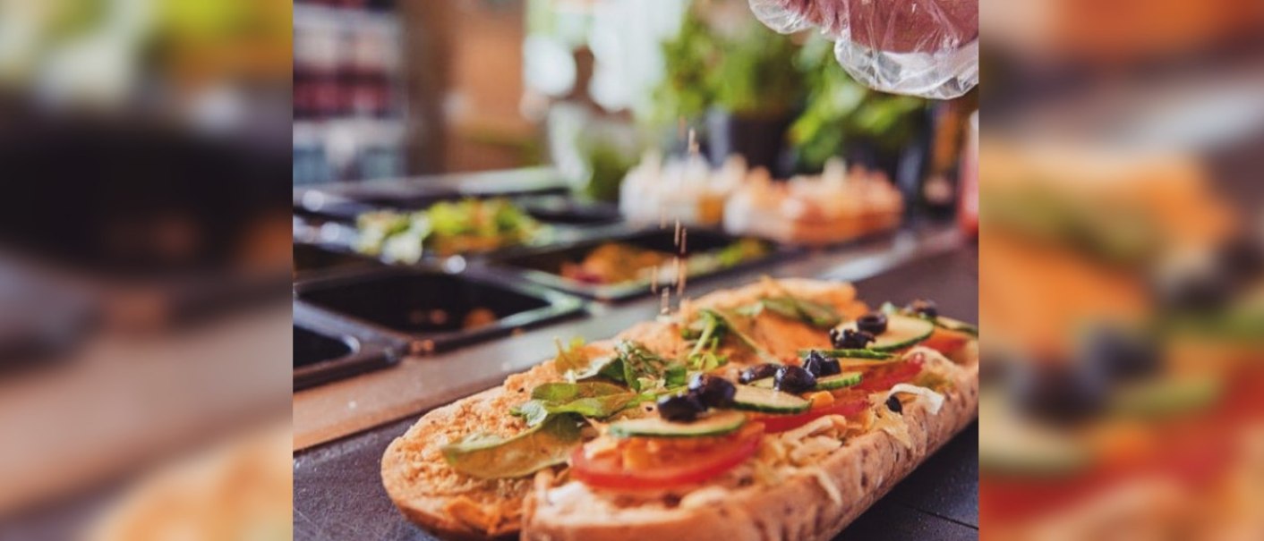 Ein Sandwich wird mit frischem Gemüse und Oliven belegt. Eine Hand streut Gewürze darüber. Im Hintergrund sind Zutaten in Behältern zu sehen., © FreshSub Ein Sandwich wird mit frischem Gemüse und Oliven belegt. Eine Hand streut Gewürze darüber. Im Hintergrund sind Zutaten in Behältern zu sehen., © FreshSub