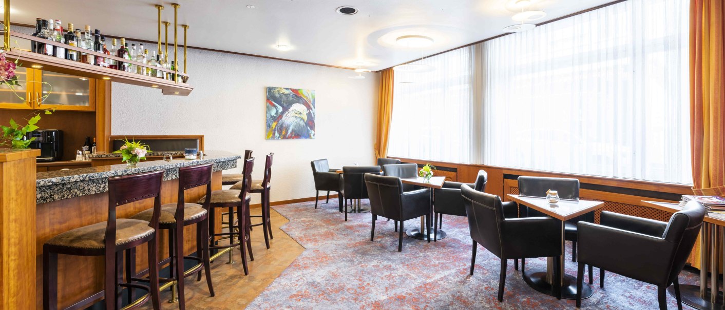 Gemütliche Hotelbar mit Theke, Barhockern und Sitzbereich. Helle Vorhänge, Teppichboden und ein buntes Gemälde an der Wand., © TOMAS