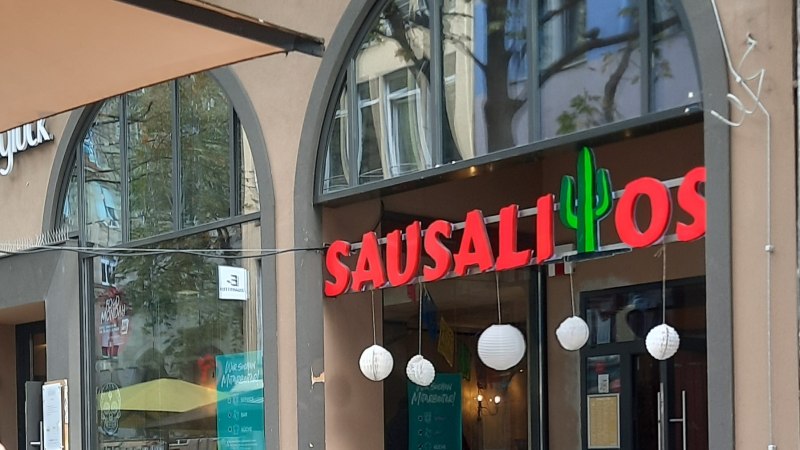 Eingang des Restaurants Sausalitos mit rotem Schriftzug und Kaktus-Dekoration. Außenbereich mit Holztischen und Werbetafel für Taco Tuesday., © SMG Eingang des Restaurants Sausalitos mit rotem Schriftzug und Kaktus-Dekoration. Außenbereich mit Holztischen und Werbetafel für Taco Tuesday., © SMG