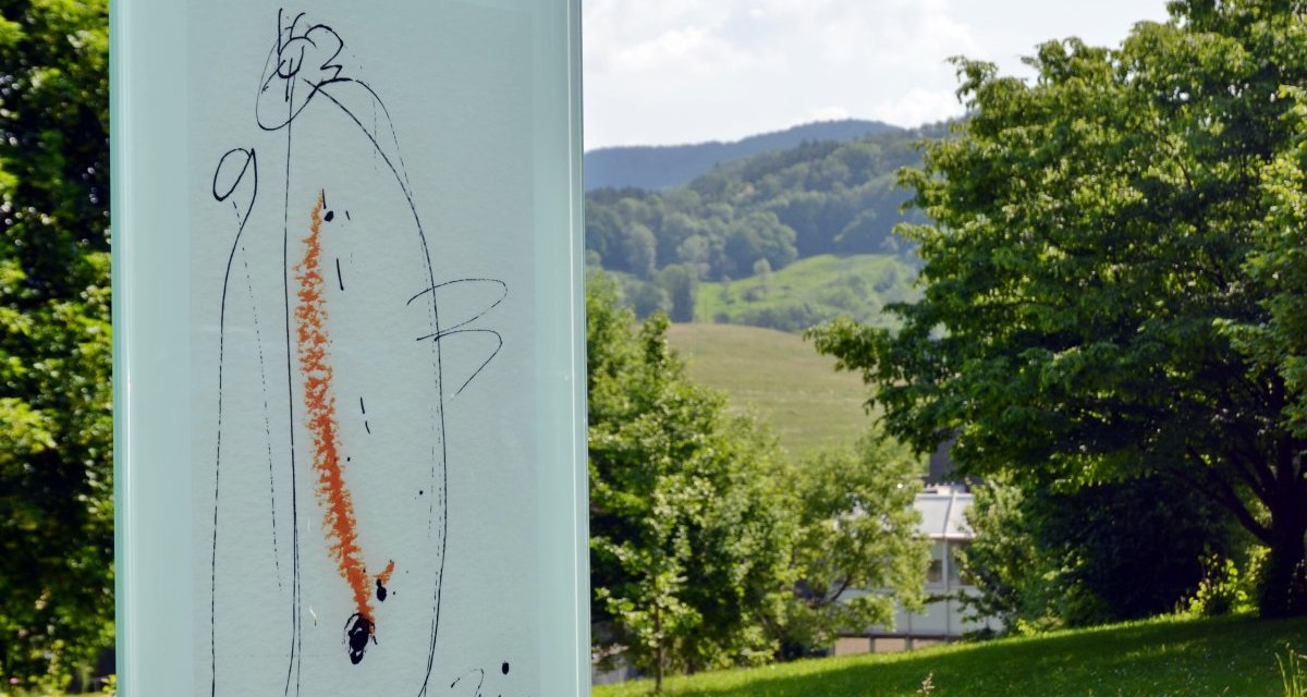 Ein Kunstwerk auf Glas zeigt abstrakte Linien und Farben. Im Hintergrund ist eine grüne Landschaft mit Bäumen und Hügeln zu sehen., © Foto: Cornelia Steinbach Ein Kunstwerk auf Glas zeigt abstrakte Linien und Farben. Im Hintergrund ist eine grüne Landschaft mit Bäumen und Hügeln zu sehen., © Foto: Cornelia Steinbach