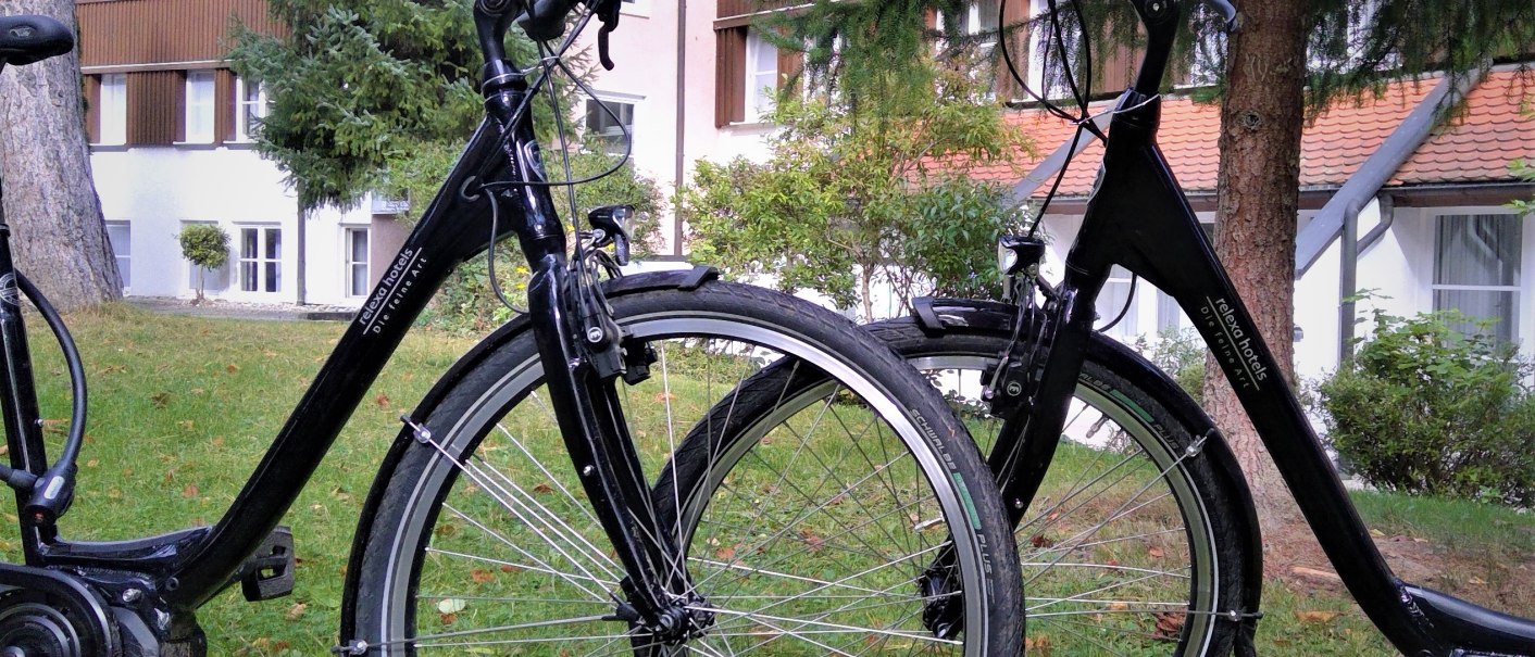 Zwei schwarze E-Bikes stehen auf einer grünen Wiese vor einem mehrstöckigen Gebäude mit Holzverkleidung und Fenstern., © relexa Waldhotel Schatten Zwei schwarze E-Bikes stehen auf einer grünen Wiese vor einem mehrstöckigen Gebäude mit Holzverkleidung und Fenstern., © relexa Waldhotel Schatten