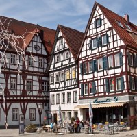 Fachwerkhäuser mit roten Dächern und blauen Fensterläden. Vor dem Wirtshaus Laurentia sitzen Menschen im Freien bei sonnigem Wetter.