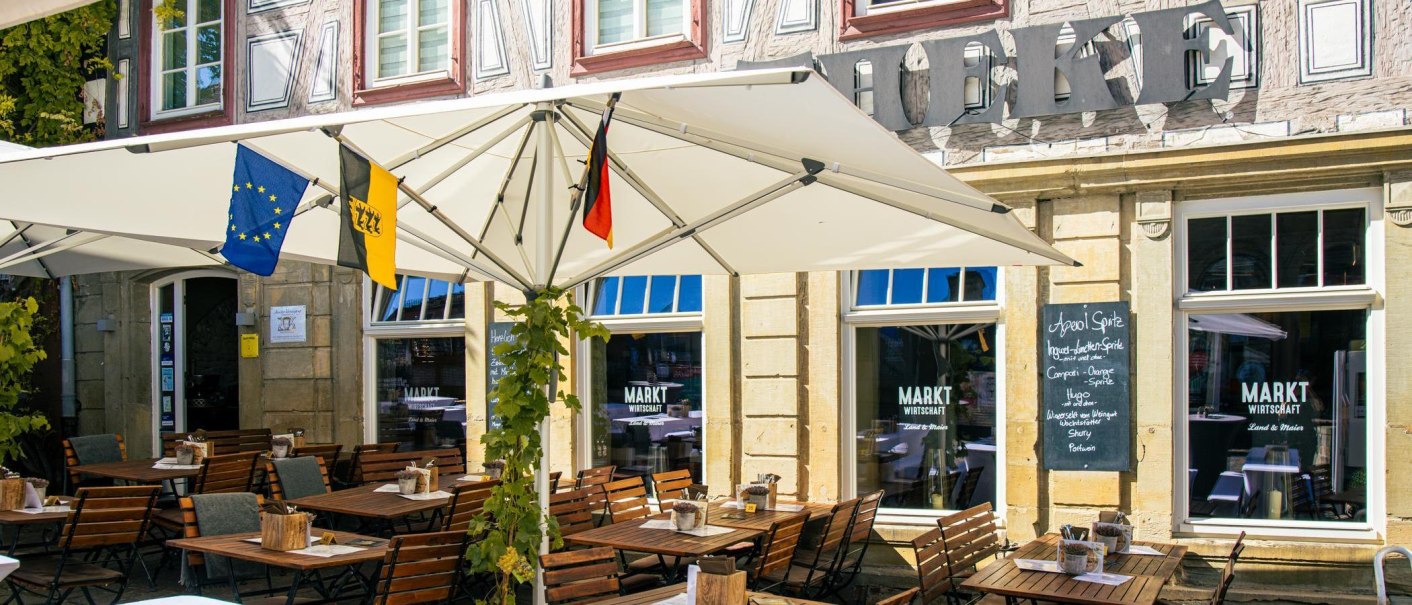 Gemütlicher Außenbereich eines Cafés in Besigheim mit Sonnenschirmen und Flaggen. Holztische und Stühle laden zum Verweilen ein., © Stuttgart-Marketing GmbH, Sarah Schmid